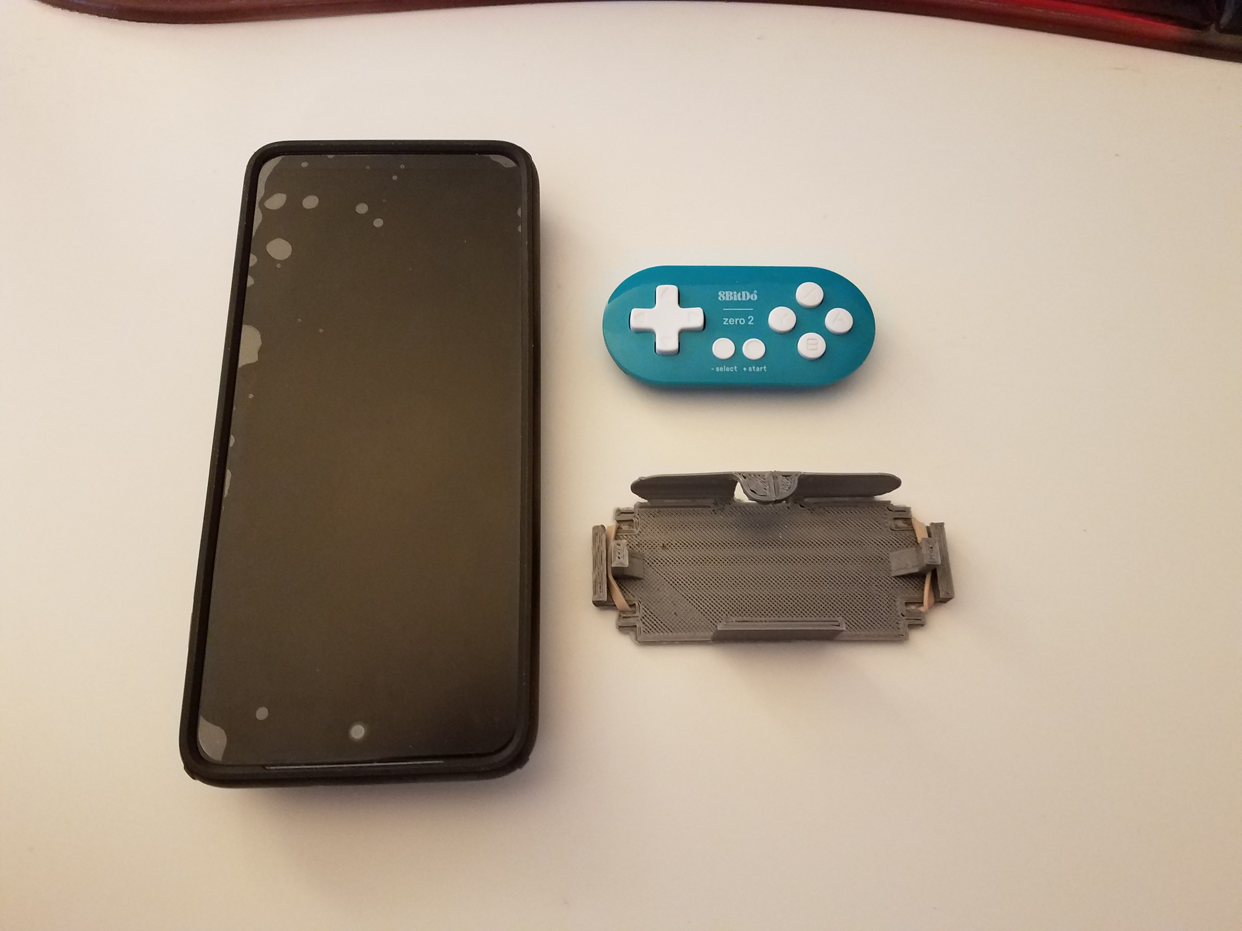 a cell phone, an 8bitdo Zero Two gamepad, and a piece of 3d printed plastic a cell phone, an 8bitdo Zero Two gamepad, and a piece of 3d printed plastic